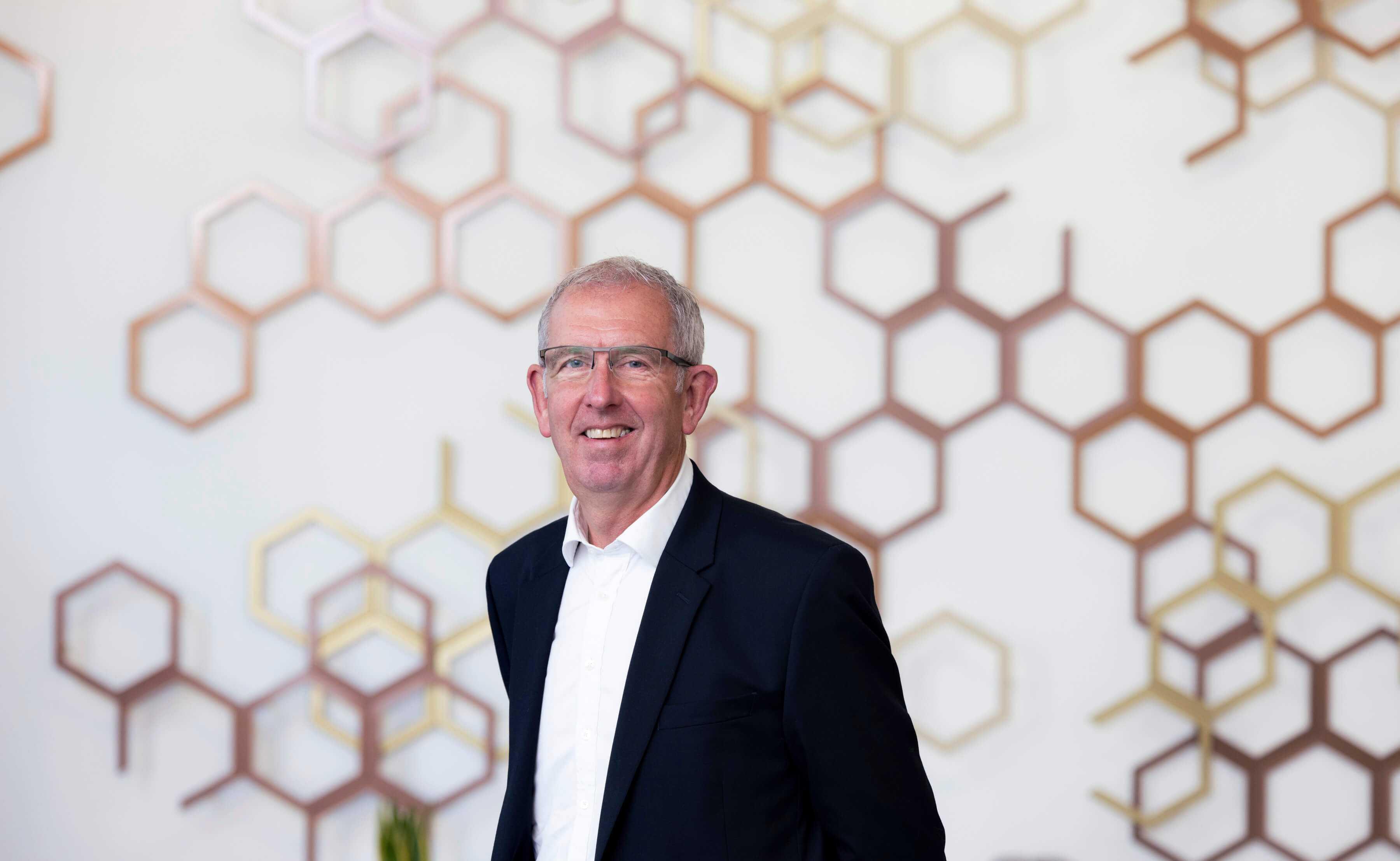 Executive Chair of InnoScot Health, Graham Watson standing in front of a white wall with bronze, hexagonal detailing across it.